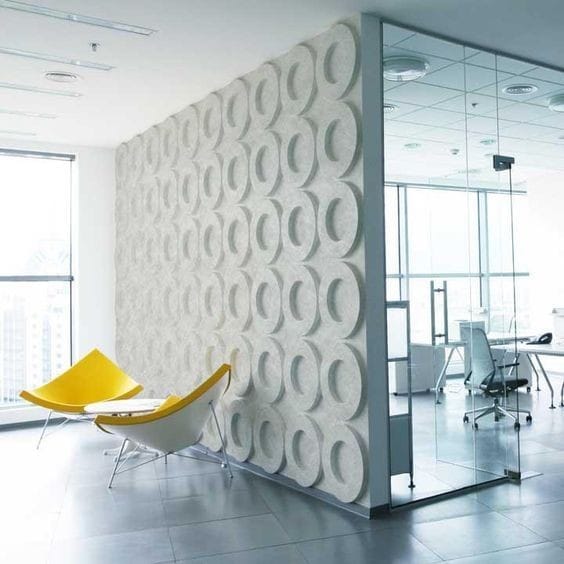 Living room with modern furniture including a gray armchair, a blue armchair, a sofa, and a round glass coffee table on a rug in a bright room with white paneled walls and large window